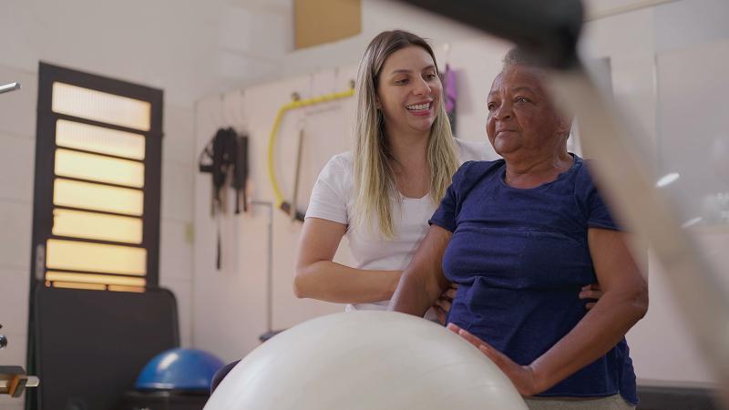 A young female physical therapist assists an older woman with balance exercises using a stability ball in a clinic setting.